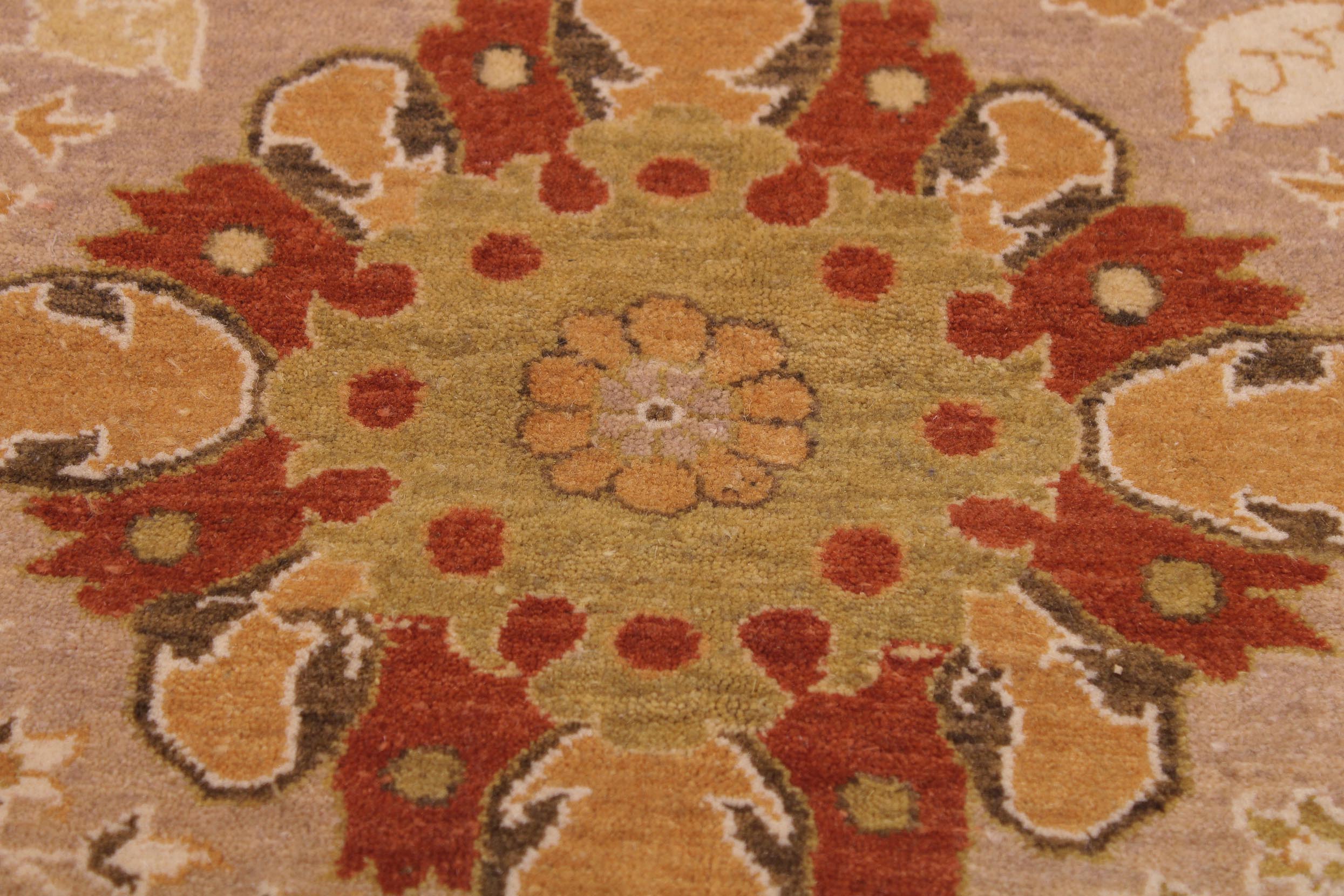 A close-up of a traditional patterned rug featuring a central floral medallion in muted yellow-green, surrounded by rust red, ochre orange, and brown motifs on a beige background.